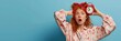 Anxious, panicking young redhead girl missed interview, overslept, holding red alarm clock and touching head frustrated with concerned face, wearing nightwear and sleep mask, blue background. 