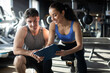 © Home-stock - Female personal trainer and her male client discussing nutrition or training plan on clipboard, sitting in modern gym interior. Healthy lifestyle, fitness concept