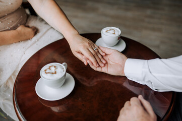 An adult man, groom and woman bride are sitting at a wooden table in a cafe holding hands with cups of milk coffee and a foam heart. Wedding photography, food, ceremony.