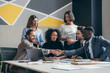 © gstockstudio - Two smiling young businessmen shaking hands while sitting with their colleagues at the office desk