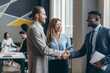 © gstockstudio - Two smiling young businessmen shaking hands in office while their colleagues working on background