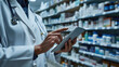 Pharmacist in a white lab coat is using a tablet in a pharmacy with shelves stocked with medications in the background.
