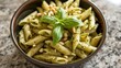  a bowl of pasta with pesto, pine nuts, and parmesan sprinkles on top of a granite countertop with a basil leaf on top.
