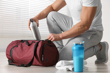 Poster - Young man putting bottle of water into bag indoors, closeup. Shaker with protein and towel on floor