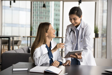 Wall Mural - Two diverse female doctor colleagues using tablet at workplace table together, talking, laughing. Happy young medical specialist woman showing electronic records to coworker