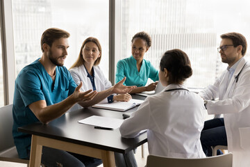 Wall Mural - Confident young surgeon man in blue medic uniform talking to multiethnic colleagues in hospital meeting room, speaking to listening diverse doctors at table, offering ideas for clinic teamwork