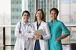 © fizkes - Cheerful diverse medical team of doctors standing together, looking at camera with toothy smiles, laughing. Happy young practitioner women in medic uniform coats posing for portrait