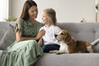 © fizkes - Happy pretty mom cuddling cute little daughter, stroking adorable dog on cozy home sofa, playing with sweet pet at home. Mother enjoying motherhood, family leisure with preschool girl and beagle