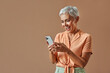 Portrait of a stylish modern beautiful gray haired middle aged woman in a pastel orange shirt and green pants holding a white phone and looking at it with joy and surprise.