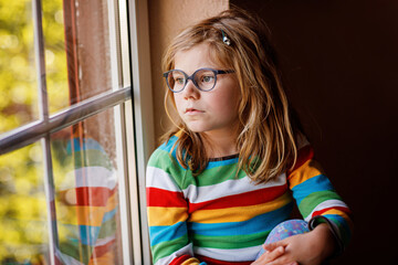 Adorable little preschool girl with eyeglasses sitting by the window. Thoughtful child looking out. Lonely kid.