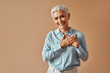 © HBS - Beautiful confident woman in blue blouse holding hands over heart while standing on beige background and smiling.