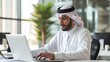 © Hope - Successful Arab Businessman in White Traditional Outfit Sitting in Office and Working on Laptop Computer. Business Manager Planning Corporate Strategy. Saudi, Emirati, Arab Businessman Concept.