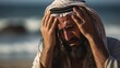 A middle aged Arab man with a beard in a keffiyeh stands on the seashore and suffers, holding his head in his hands
