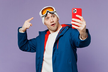 Wall Mural - Skier man wear warm blue windbreaker jacket point finger on ski goggles mask hat do selfie shot mobile cell phone spend extreme weekend winter season in mountains isolated on plain purple background.