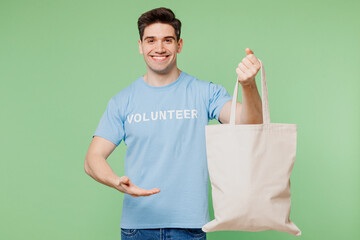 Sticker - Young man wear blue t-shirt white title volunteer hold in hand point on eco cotton textile shopper isolated on plain pastel green background. Voluntary free work assistance help charity grace concept