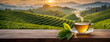 A steaming cup of tea with green leaves on a wooden surface with a scenic tea plantation at sunrise. Panorama with copy space. Banner.