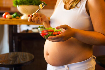 Wall Mural - Young pregnant woman eating fresh vegetable salad at the kitchen