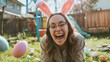 A candid shot of a woman in bunny ears laughing while hiding Easter eggs in a backyard