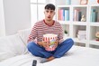 Young hispanic man eating popcorn sitting on the bed watching a movie in shock face, looking skeptical and sarcastic, surprised with open mouth