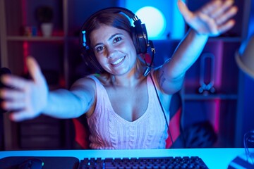 Wall Mural - Young blonde woman playing video games wearing headphones looking at the camera smiling with open arms for hug. cheerful expression embracing happiness.