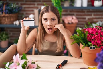 Poster - Young blonde woman working at florist shop holding credit card scared and amazed with open mouth for surprise, disbelief face