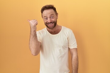 Wall Mural - Middle age man with beard standing over yellow background with a big smile on face, pointing with hand finger to the side looking at the camera.