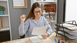 © Krakenimages.com - A joyful woman with glasses celebrates success while working with documents in a modern office.