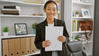 A professional chinese woman smiling, holding documents in a modern office setting with shelving and decor.