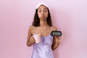Wall Mural - Young hispanic woman wearing nightgown holding alarm clock puffing cheeks with funny face. mouth inflated with air, catching air.