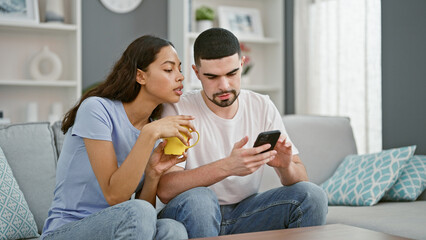 Wall Mural - Beautiful couple cozily sitting together on their sofa at home, immersed in their smartphone screen, sipping coffee, lost in their online world.