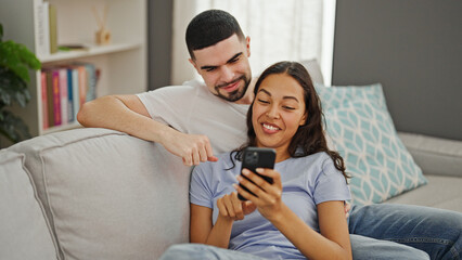 Poster - Beautiful couple sitting on a comfy sofa, smiling together enjoying a fun smartphone moment in their cozy living room