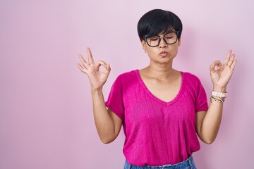 Poster - Young asian woman with short hair standing over pink background relaxed and smiling with eyes closed doing meditation gesture with fingers. yoga concept.