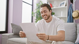 Fototapeta Panele - Young hispanic man reading document sitting on sofa smiling at home