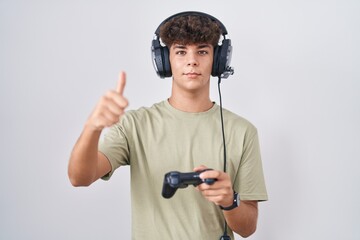 Wall Mural - Hispanic teenager playing video game holding controller approving doing positive gesture with hand, thumbs up smiling and happy for success. winner gesture.