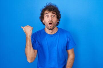 Wall Mural - Hispanic young man standing over blue background surprised pointing with hand finger to the side, open mouth amazed expression.