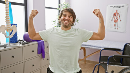 A cheerful young man flexing muscles joyfully in a well-equipped rehabilitation clinic's therapy room.