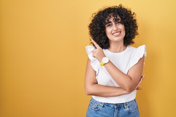 Poster - Young middle east woman standing over yellow background with a big smile on face, pointing with hand and finger to the side looking at the camera.