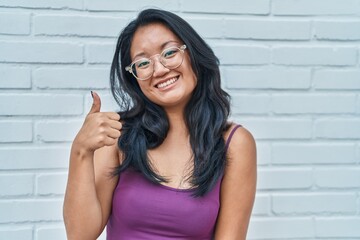 Canvas Print - Asian young woman standing over bricks background smiling happy and positive, thumb up doing excellent and approval sign