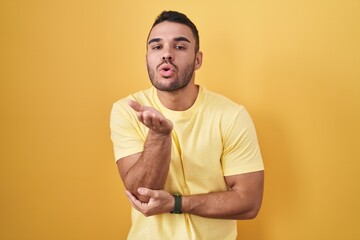 Poster - Young hispanic man standing over yellow background looking at the camera blowing a kiss with hand on air being lovely and sexy. love expression.