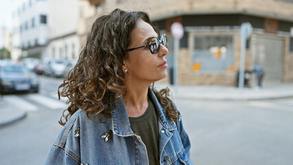 Sticker - A middle-aged hispanic woman with curly hair wearing sunglasses stands thoughtfully on a sunny urban street.