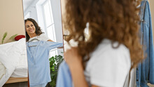 Smiling woman with curly hair trying on blue shirt in front of mirror inside her light bedroom