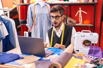 Canvas Print - Young hispanic man with tattoos dressmaker designer using laptop at tailor room scared and amazed with open mouth for surprise, disbelief face