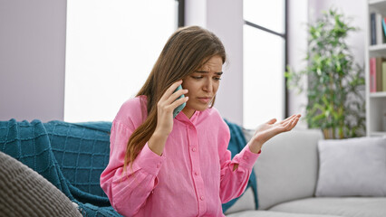 Unhappy, worried young beautiful hispanic woman talking with serious problems via smartphone, sitting alone at home on the living room sofa.