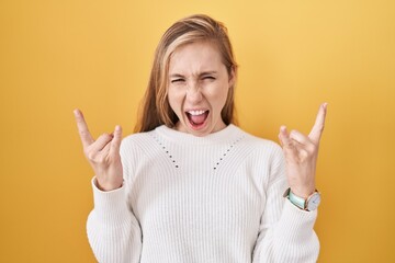 Young caucasian woman wearing white sweater over yellow background shouting with crazy expression doing rock symbol with hands up. music star. heavy music concept.