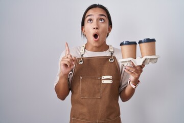 Sticker - Young hispanic woman wearing professional waitress apron holding coffee amazed and surprised looking up and pointing with fingers and raised arms.