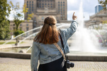 Cheerful Young Woman Walking Near Palace Of Culture And Science In Warsaw City, Poland. Redhead Girl With Camera Pointing Finger Outdoors. Traveling In Summer. Urban Lifestyle Concept. Rear View