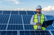 © Alan - portrait of an engineer smiling with confidence using a laptop in front of a solar panel power station, handsome male technician electrician looking at camera holding a computer