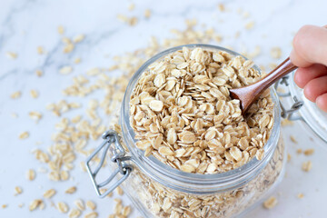 Rolled oats (oatmeal flakes) in a glass jar with hand holding a wooden spoon. Top table view. Organic healthy cereal food for breakfast. Selective focus.