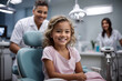 Little girl smilimg in a dentist chair in clinic after dental checkup or treatment. Visiting dentist with children. Doctor dentist on the background.
