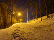 Park wieczorową porą, śnieg, latarnie oraz stary mur.